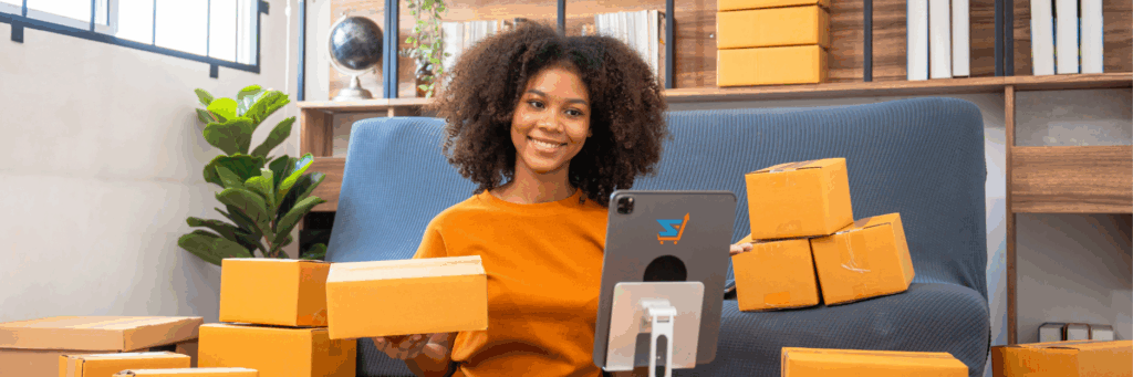 Smiling woman sitting on a sofa surrounded by cardboard boxes while managing her Amazon business on a tablet.
